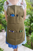 Green canvas gardening apron with assorted pockets containing gardening tools modelled by a woman.