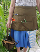 Woman wearing a half gardening apron made of green canvas. The many pockets contain tools, scissors and phone. She is holding a gardening tool basket