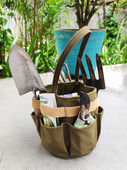 Green canvas gardening tool basket containing various garden tools and a seed packet