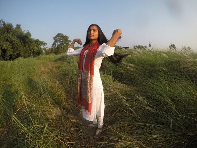 A woman wearing a rust coloured scarf with a border of mottled colour weave. She is stood in a field with one hand up