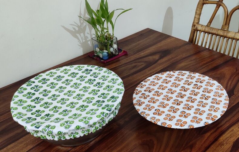 Two round bowls on a wooden table. The bowls are covered with printed, organic cotton covers. One is white with orange flower porint, the other i d ehite with green fliwer print.
