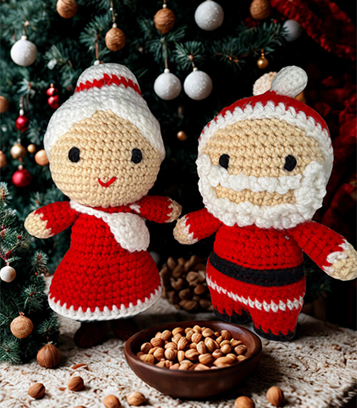Two crochet Christmas dolls propped up in front of a decorated Christmas tree with a bowl of hazelnuts in front of them