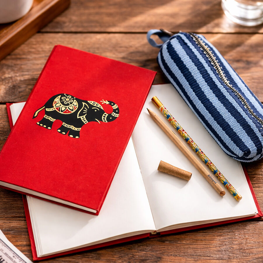 Note book with a bright red cover and a print of a black and gold elephant. There is another note book open with a hand painted pencil and a paper pen resting on the page and a blue striped pencil case