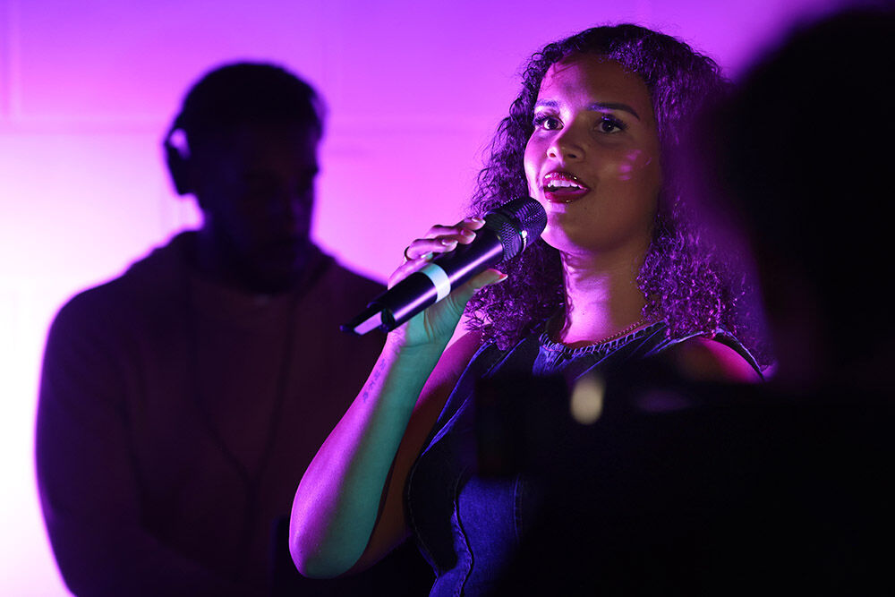 Photo of a young woman singer, performing with a DJ behind, in purple lighting