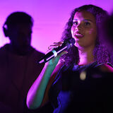 Photo of a young woman singer, performing with a DJ behind, in purple lighting