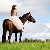 A portrait of a young woman, wearing an elegant long dress, sitting on a chestnut horse, with blue sky and clouds behind.
