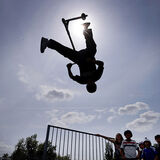 A scooter rider flips upside down on a skatepark ramp, silhouetted against the sky, with a groups of kids looking on