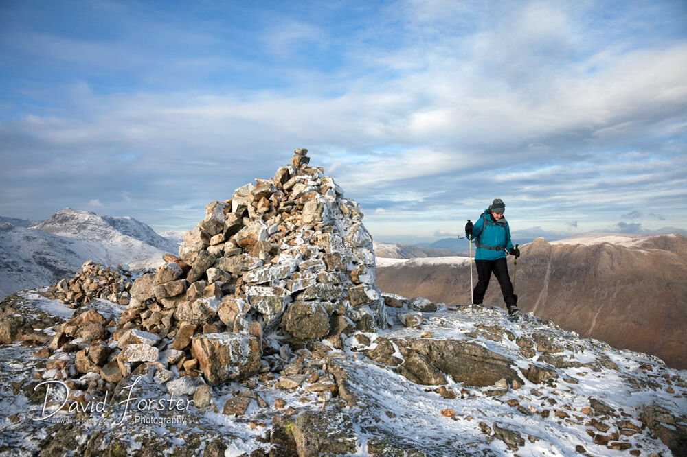 05D-9933 Walker on the Summit of Pike of Blisco in Winter with the Mountain of Bow Fell to the Left of the Cairn Lake District Cumbria UK