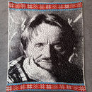 knitted portrait of a white man resting his chin on his hand. top and bottom borders are red with white and blue with yellow Fair Isle style stripes.