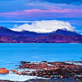 A coastal sunset view looking towrds Gruinard bay in the Scottish Highlands showing rugged rocks in the foreground and pink clouds above distant snowy mountains.