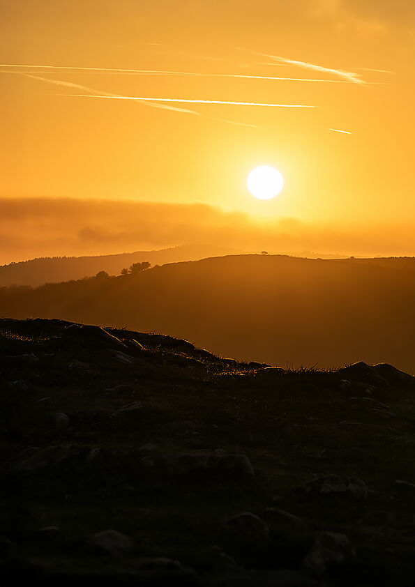 Silhouette of rolling hills at dawn under a bright sun with clear contrails in an orange sky.