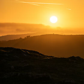 Silhouette of rolling hills at dawn under a bright sun with clear contrails in an orange sky.