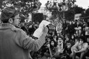a young man reads out a poem to the group of young people sitting in a park