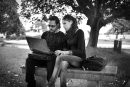 young man and woman sit at the park bench with an open laptop